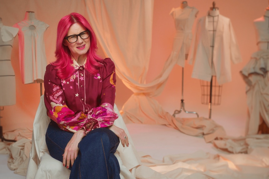 Glynis, with bright pink shoulder-length hair, sits on a chair in front of fashion bodices and fabric