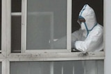 A person wearing a white biohazard suit leans on a window of a high-rise building.