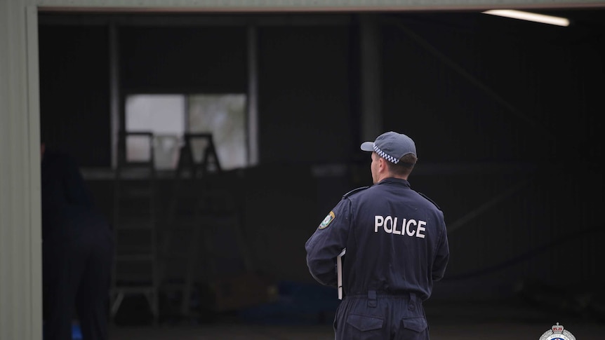 A policeman in a jumpsuit looks into an empty, dark shed.