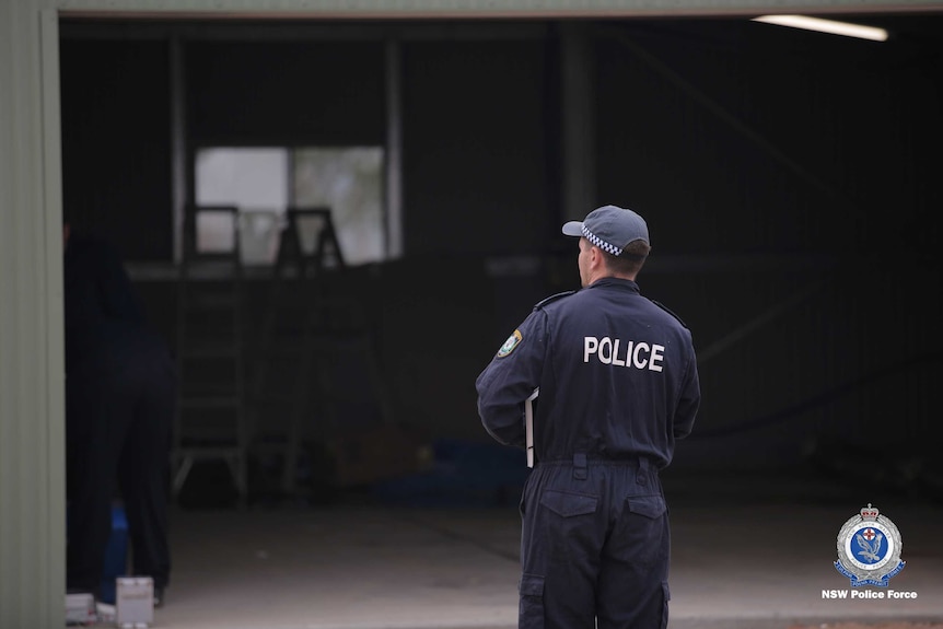 A policeman in a jumpsuit looks into an empty, dark shed.