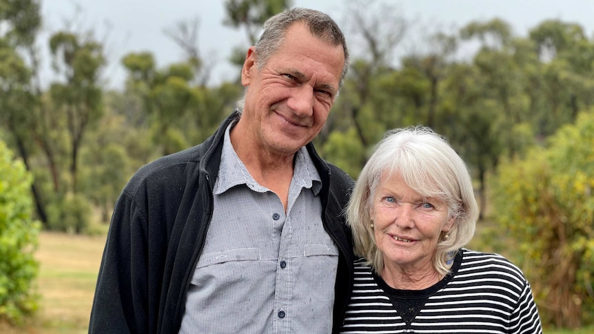 An elderly couple smiles as they stand on their property. The trees behind them have green leaves but the trunks remain burnt.