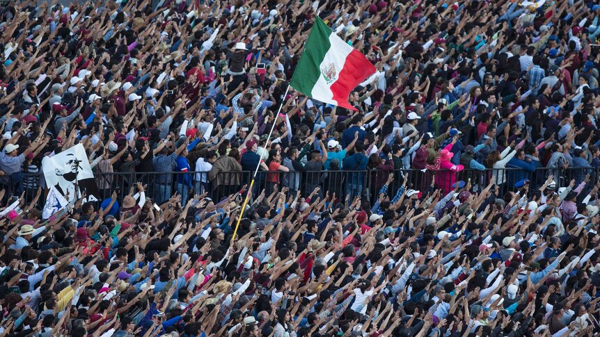 A large crowd stands with their hands in the air and a waving Mexico flag.