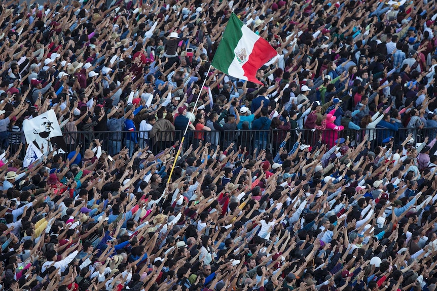 A large crowd stands with their hands in the air and a waving Mexico flag.
