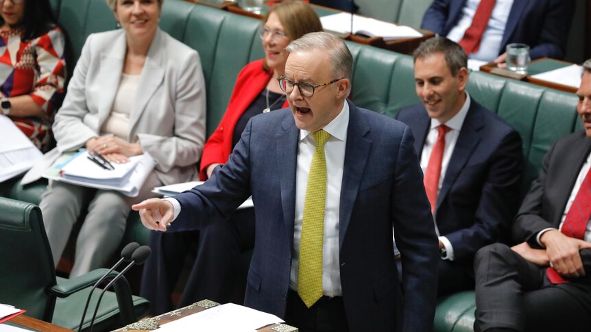 A middle-aged white man in a suit with a yellow tie and glasses yells and points while speaking in parliament.