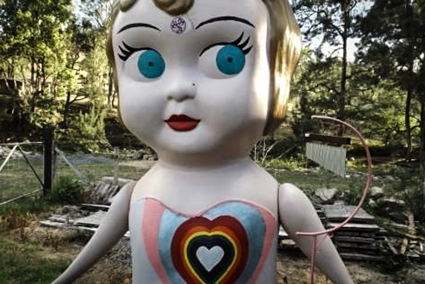 Giant kewpie doll painted with rainbow heart, with bushland in background