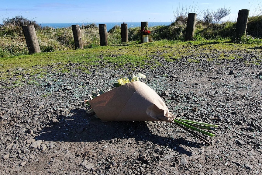 Flowers lie on the ground in a gravel car park next to the ocean.