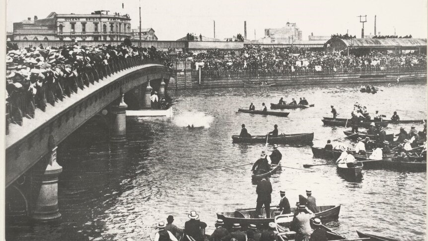 Harry Houdini splashes into the Yarra River as crowds watch on from the bank, a bridge and boats.