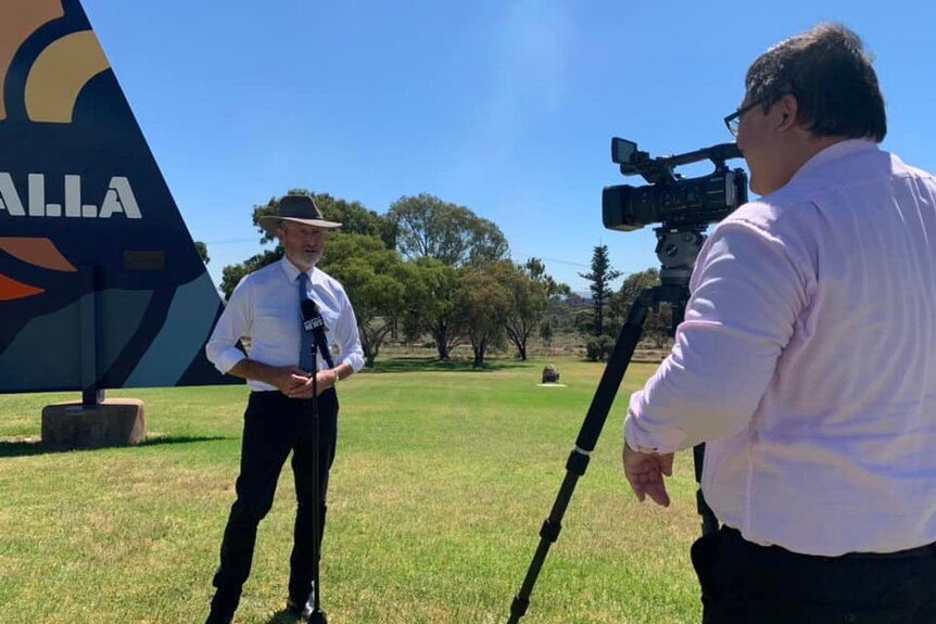 A man in an akubra hat stands in front of a video camera