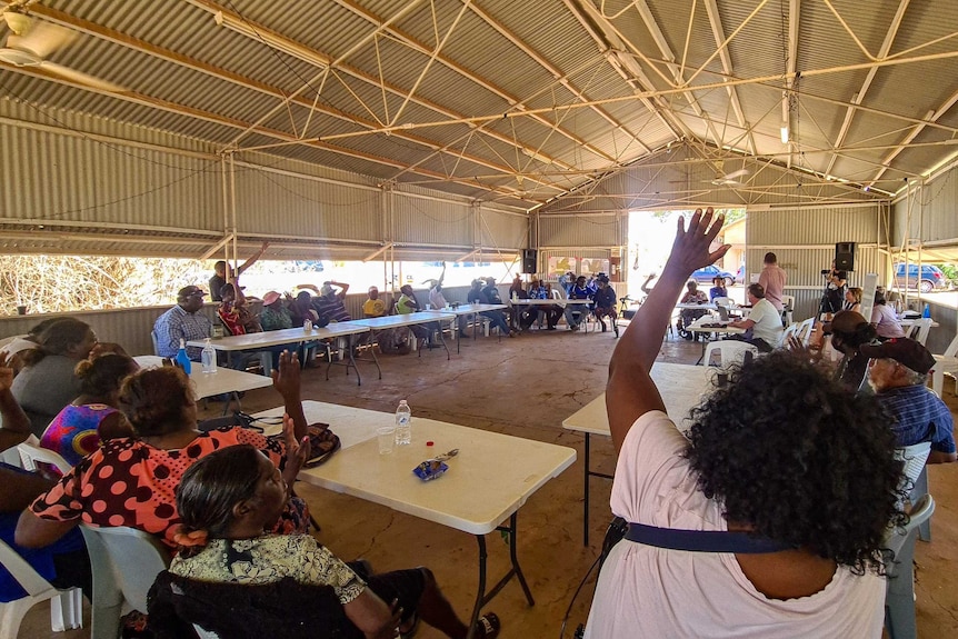 A group of Indigenous traditional owners raise their hands at a meeting to vote, they are in a large correlated iron room.