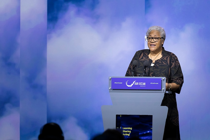 Fiame Naomi Mata'afa stands behind a lectern in front of a blue-purple cloudy backdrop. 