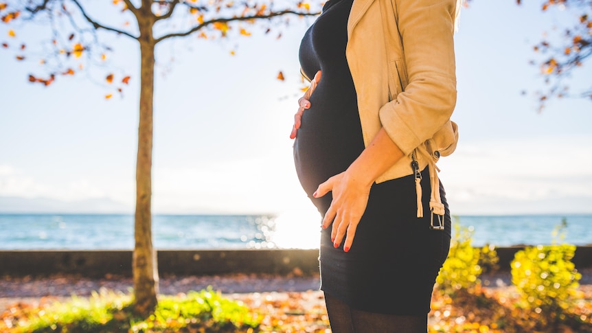 Woman stands near water holding her early pregnant stomach in story about when everyone is excited about your pregnancy but you