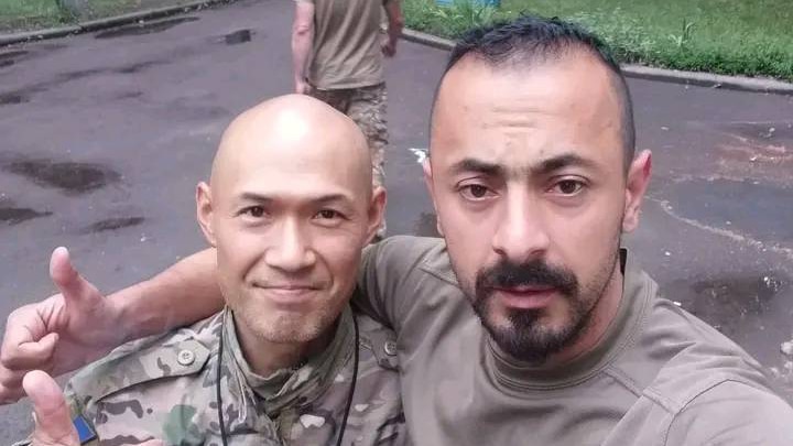 Two men in military gear gather together for a selfie in front of a burnt-out car