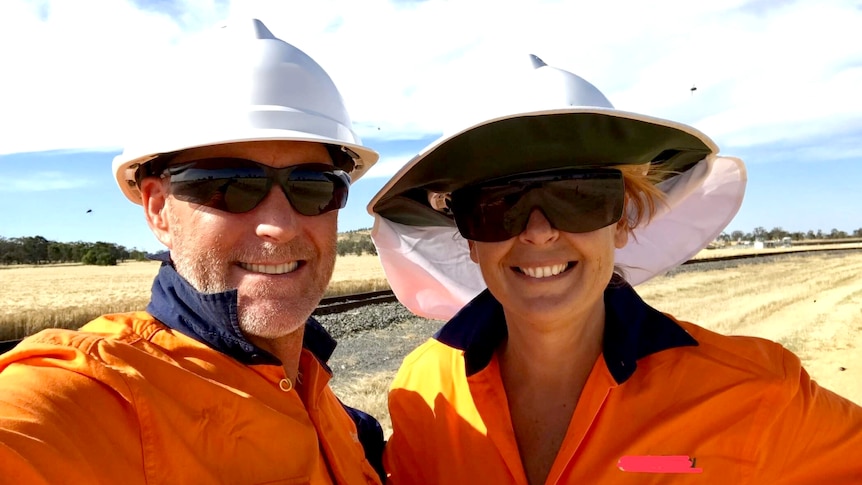 A man and a woman in high-vis, helmets and dark sunglasses smile for a selfie, a paddock behind them.