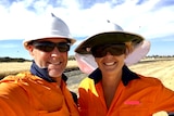 A man and a woman in high-vis, helmets and dark sunglasses smile for a selfie, a paddock behind them.