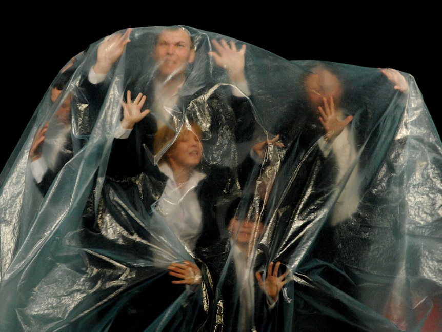 A small group of people wearing suits, inside a thick plastic bag, trying to push through it, looking worried.