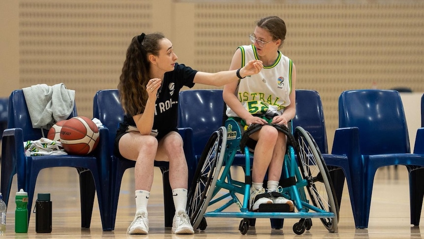A woman basketball coach sits on a chair and gives instructions to a young woman wheelchair basketballer.