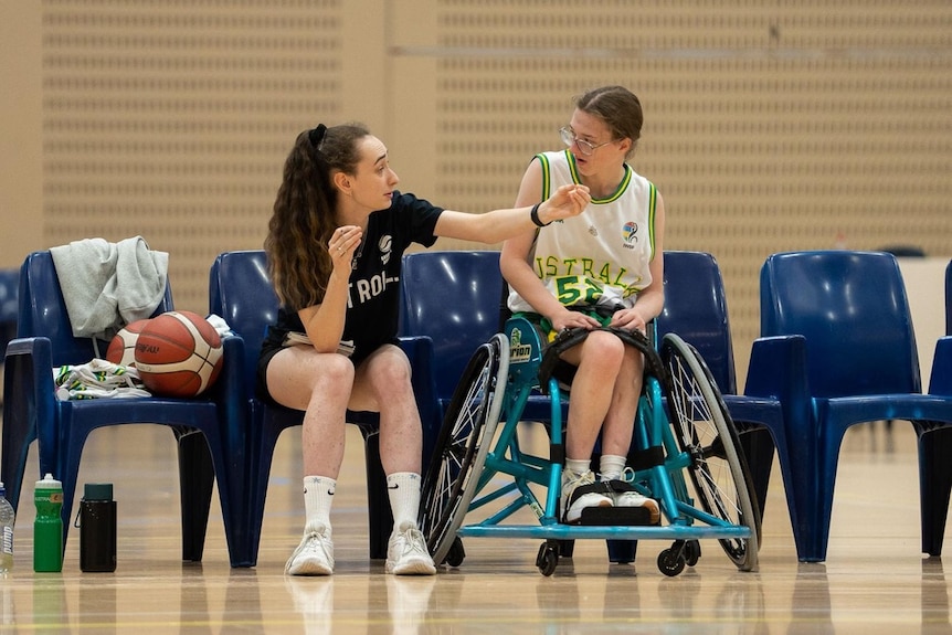 A woman basketball coach sits on a chair and gives instructions to a young woman wheelchair basketballer.