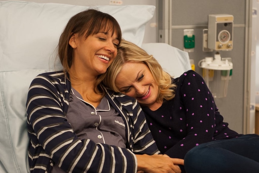 Parks and Recreation characters Leslie and Ann snuggle up in a hospital bed.