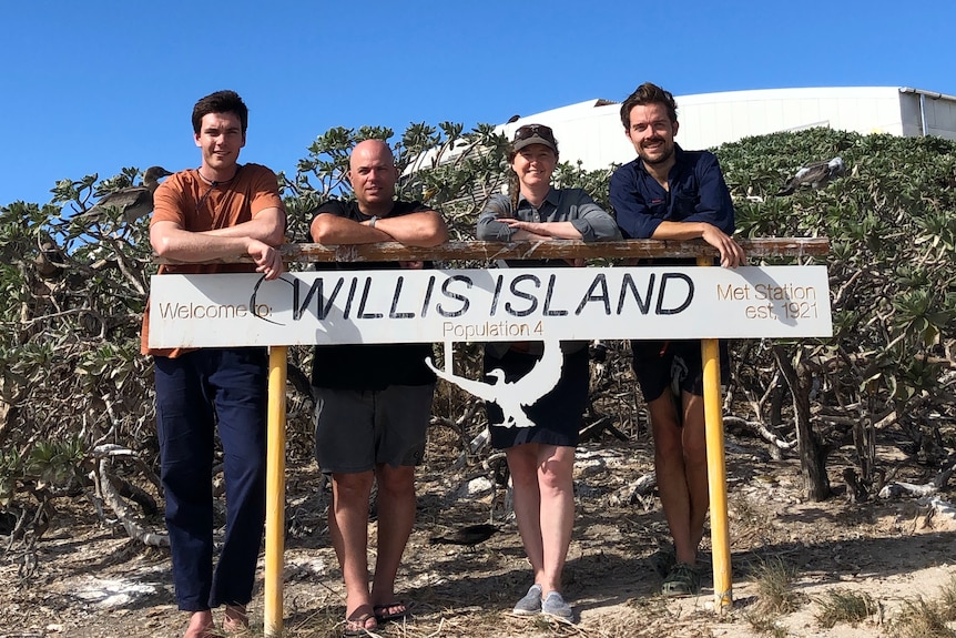 Three men and a woman lean on a sign that reads 'Willis Island. Population: 4'