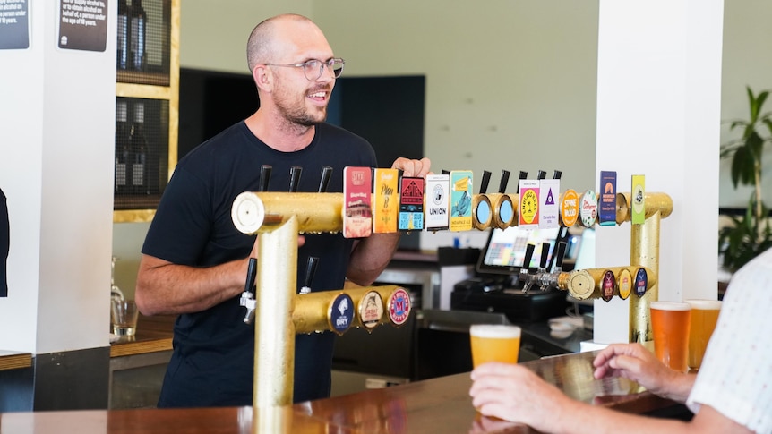 A tall man standing behind the beer taps at a bar smiling at customers.