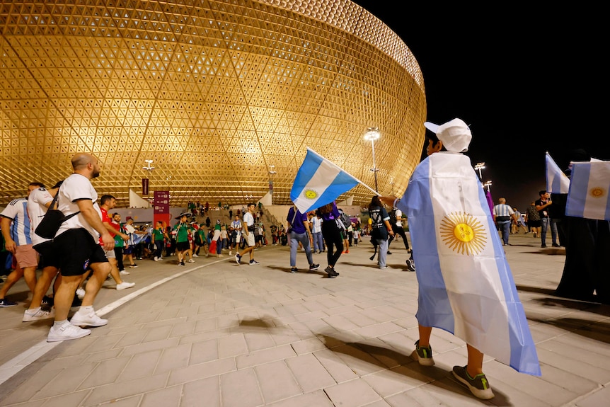 A person wrapped in an Argentina flag stands outside a stadium as other people walk past. 