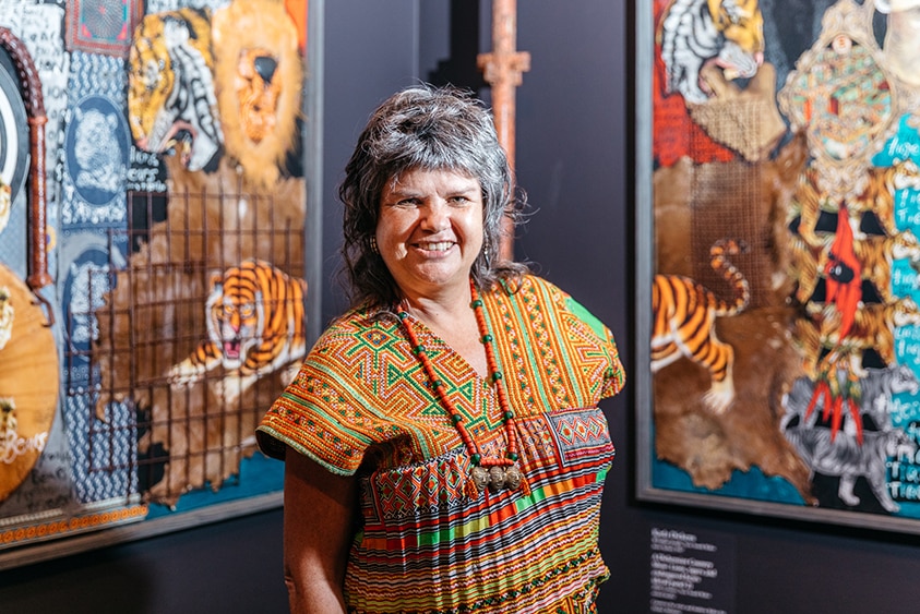 A smiling woman with short grey hair wears bright patterned top and stands in gallery with bright mixed media artworks on wall.