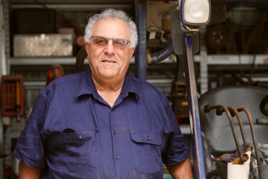 a man in a blue work shirt stands in front of tools in the shed smiling at the camera