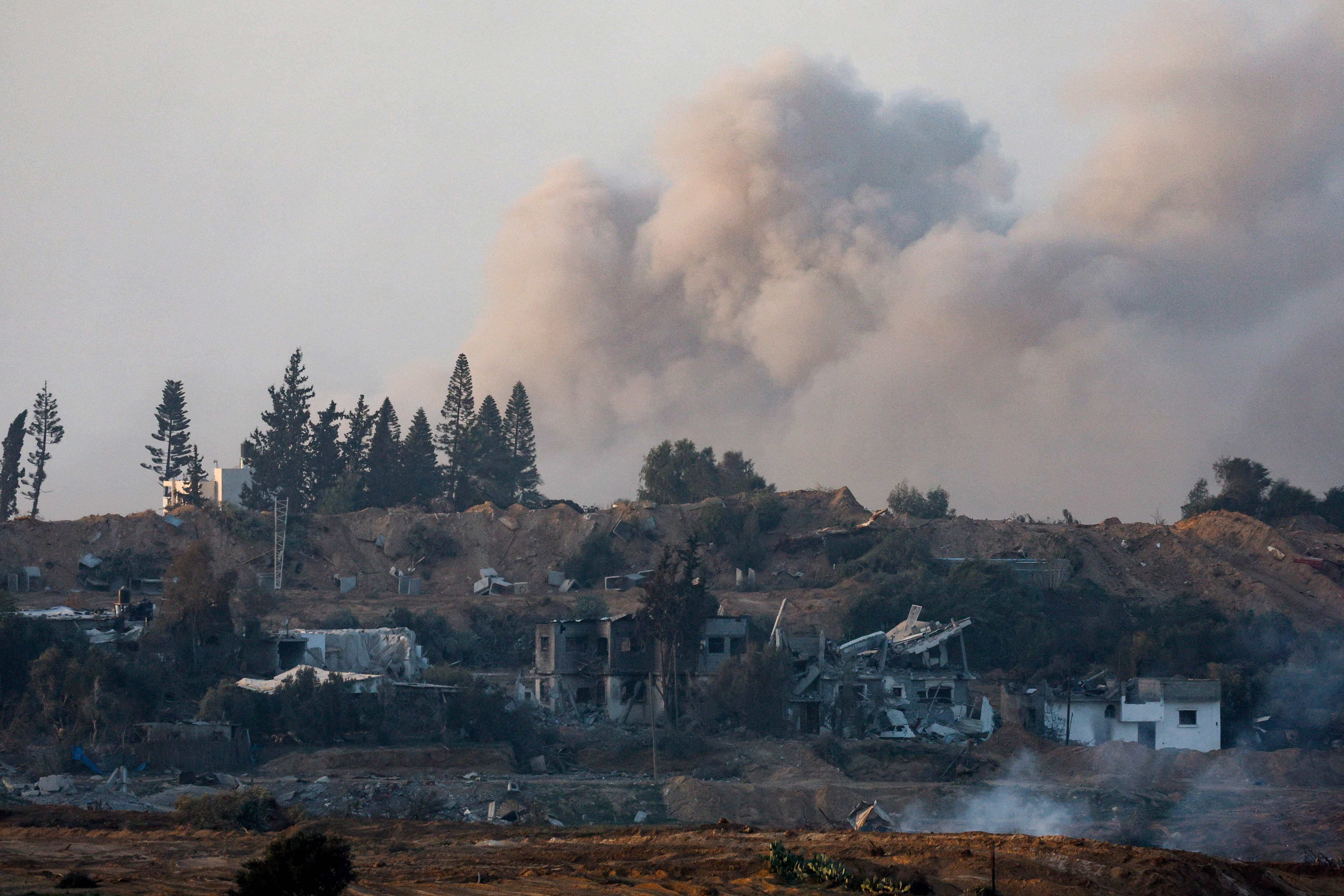 Smoke rises during an explosion with severely damaged buildings seen near a low hill.