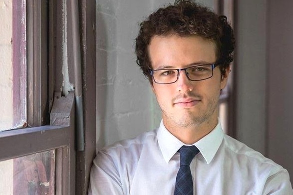 A man wearing glasses with curly black hair stands near a window looking at the camera.