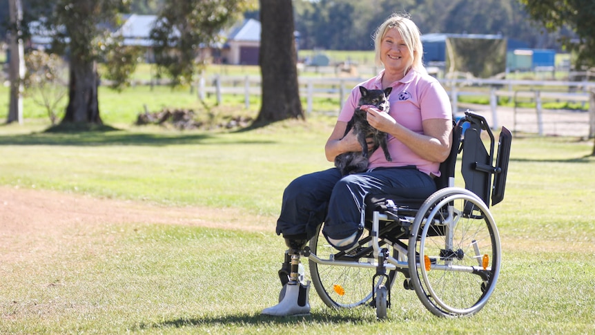 A woman with two leg amputations sits on a wheelchair smiling and holding her small dog.