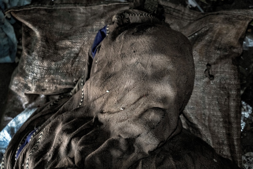 The outline of a face is faintly visible through a heavy brown shawl covering the head of a dead body.
