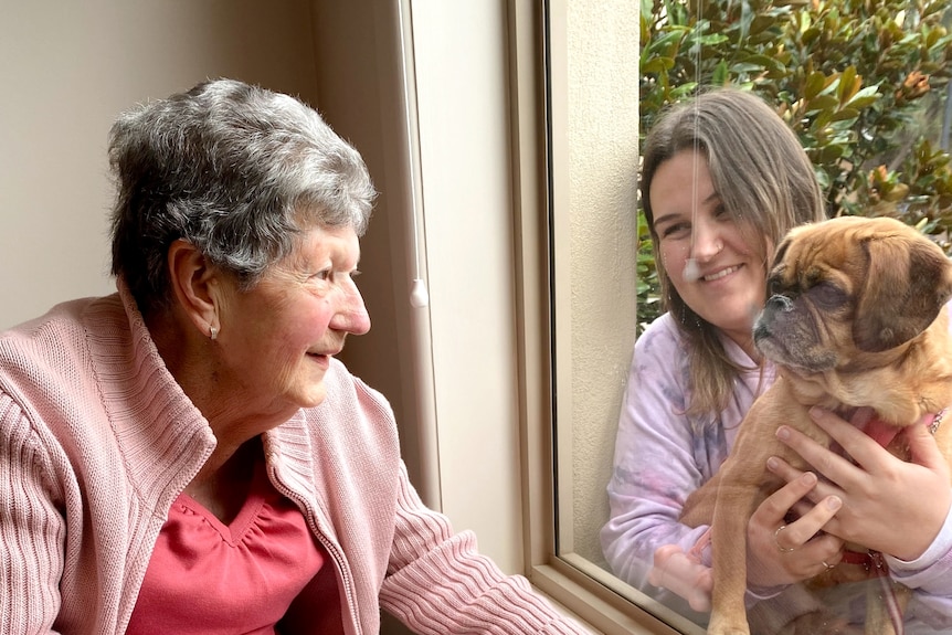 A photo of Nancye smiling and looking through a window with her grandchild and a dog on the other side