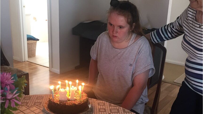 A young woman with a pony tail, wearing a grey t-shirt, sits at a kitchen table with a birthday cake in front of her