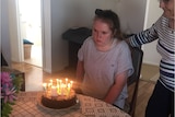 A young woman with a pony tail, wearing a grey t-shirt, sits at a kitchen table with a birthday cake in front of her