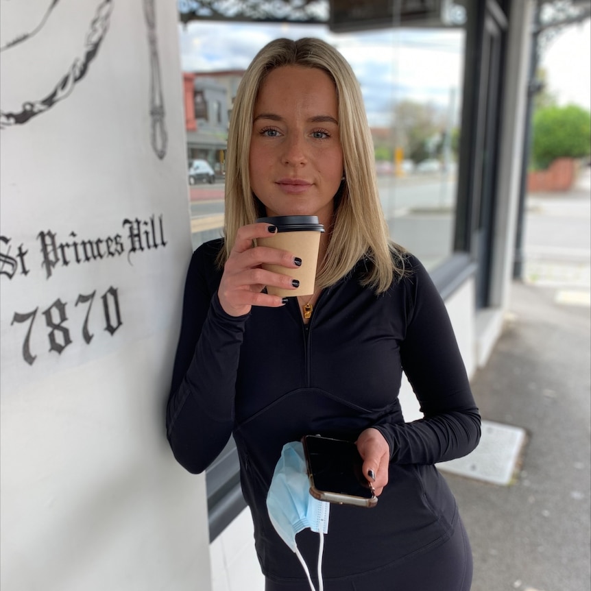 A blonde woman holds a takeaway coffee cup outside a cafe. She's wearing a black top.