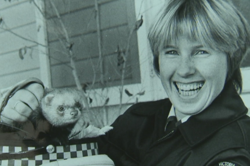 A black and white photo of a female police officer, holding a ferret in her hat.