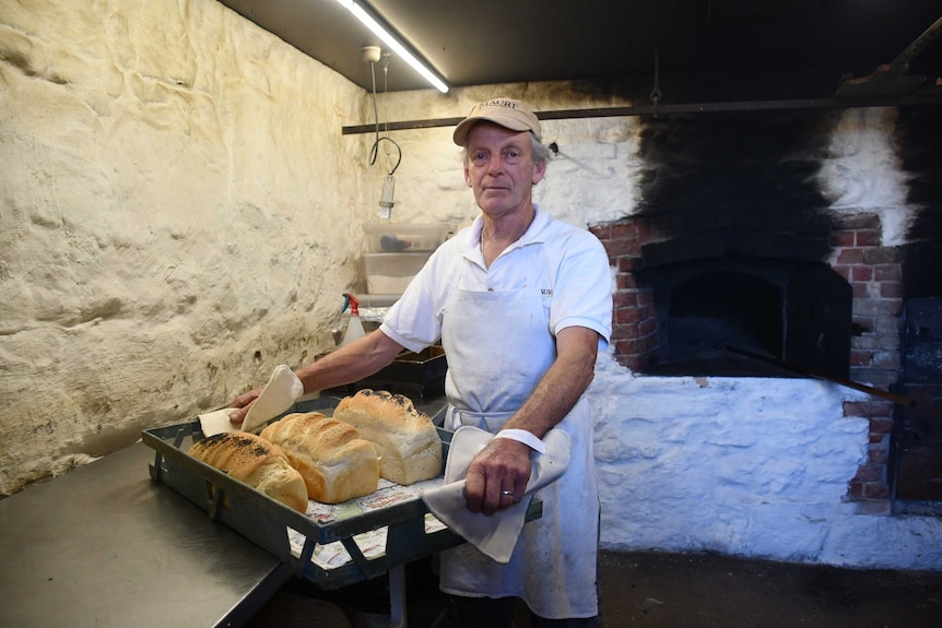 A man in a white baker's uniform wearing a cream-coloured cap stands in an underground bakery holding a batch of fresh bread.