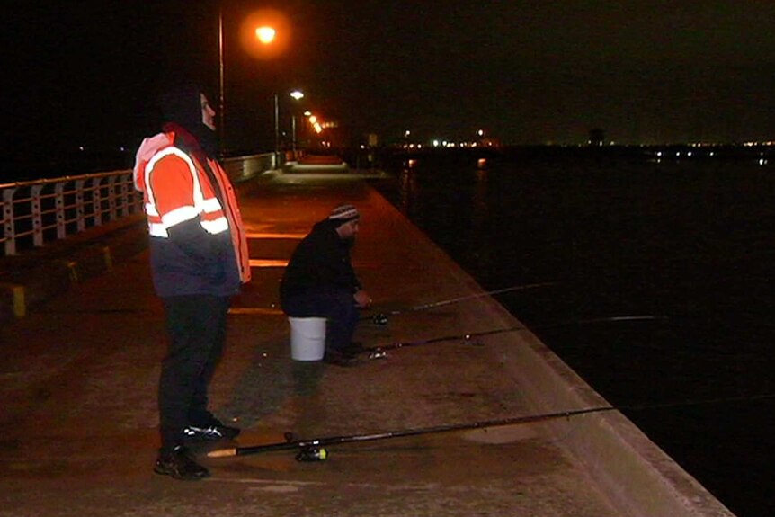 A man in a high-vis vest and another sitting on an upturned bucket at St Kilda Pier at night.