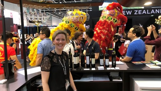 A woman sits at a busy wine stall, there are yellow and red chinese dragons and a crowd in the background