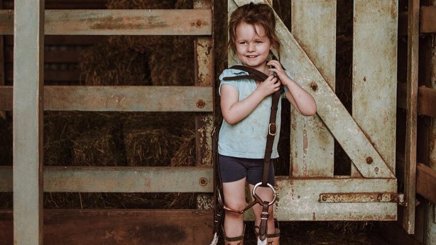 A young girl stands in an old farm shed, holding a bridle.