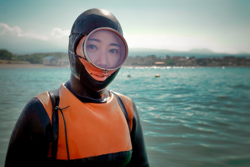 A woman wearing a full body diving suit and a mask smiles while standing infront of the sea.