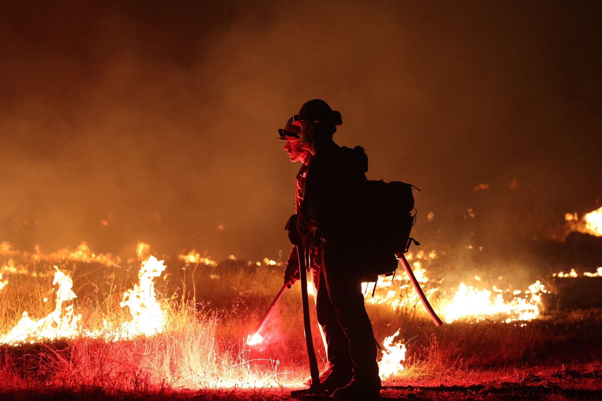 Two fire fighters stand in emergency gear surrounded by brush fires. 