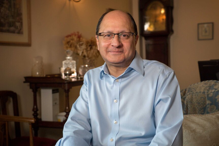 A balding man wearing glasses and a blue shirt sits on a couch.