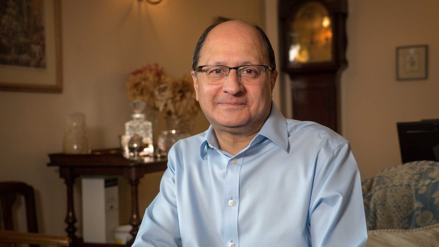 A balding man wearing glasses and a blue shirt sits on a couch.