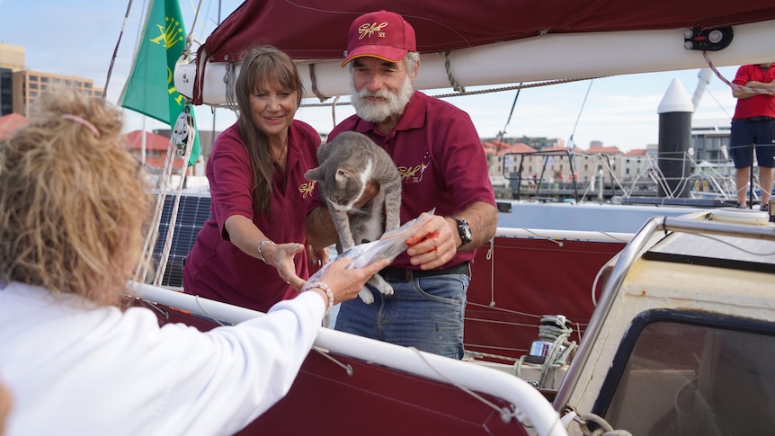 A man with a white beard in a maroon uniform holds a cat and accepts a clear bag of cat treats.