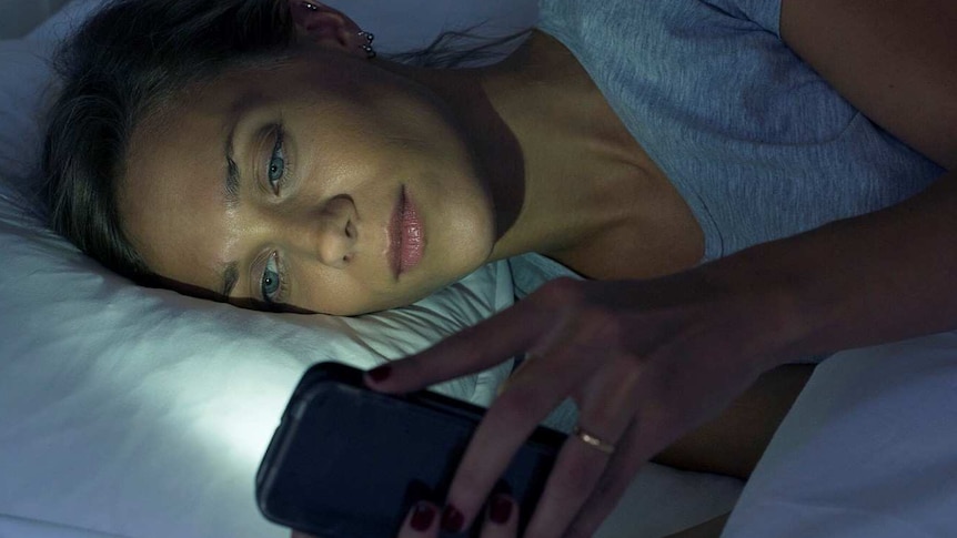 Woman lying on her side in bed using mobile phone with light from screen reflected on her face