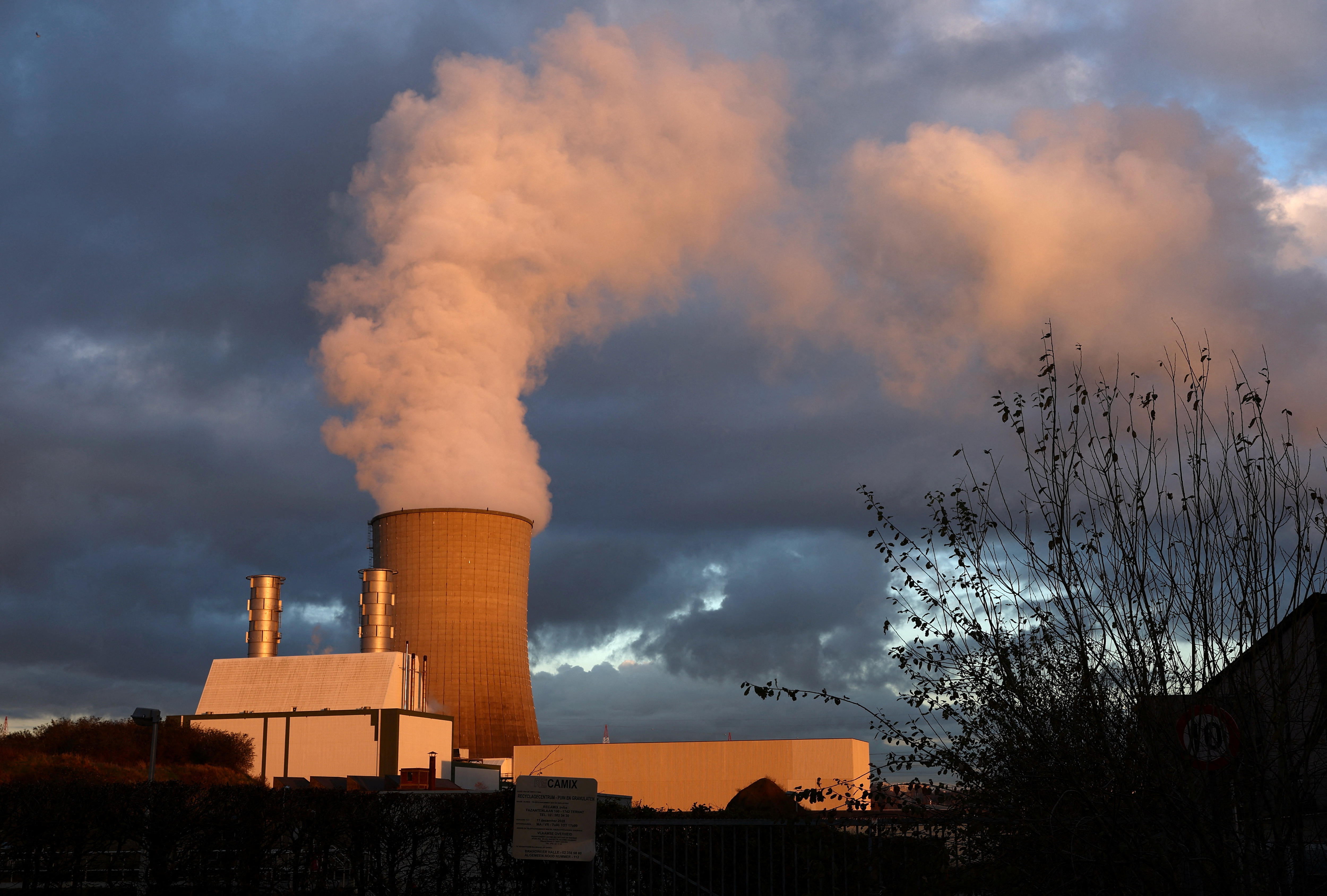 Smoke billows out of a power plant during golden hour on a cloudy day, casting the scene in an orange hue.