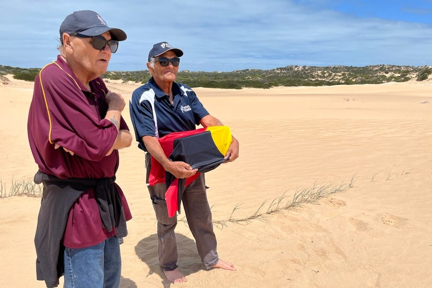 Two aboriginal men holding a box and standing on sand.