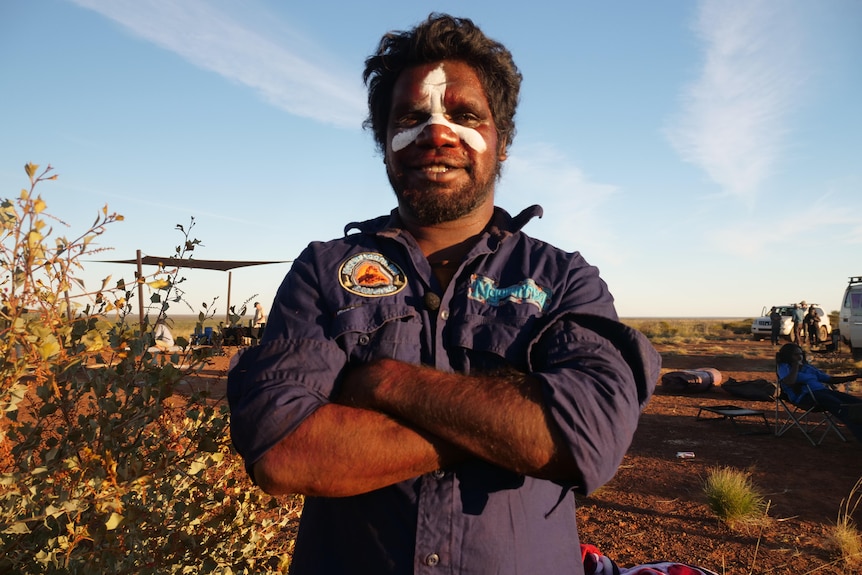 Indigenous man stands with his face painted and arms folded