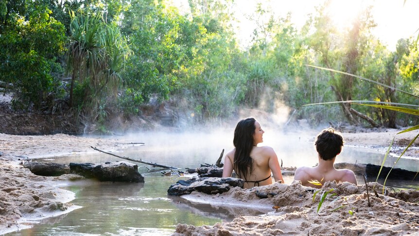 Two people sit in hot springs water surrounded by steam and sand